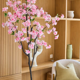 De Kunst Kersenbloesem 150 cm roze, met zijn delicate roze zijden bloesems, staat elegant in een moderne kamer met houten planken, decoratieve voorwerpen, een beige stoel en een groen kussen – en voegt zo de charme van zijden bloemen toe aan je interieur.