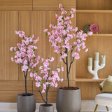 Drie Kunst Kersenbloesem-bomen van 150 cm met roze bloesems staan voor een houten lambrisering, naast planken die versierd zijn met decoraties en een moderne witte kandelaar.