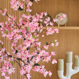 Een kunstplant bougainvillea van 150 cm, roze van kleur en met roze bloesems, staat voor houten planken waarop een decoratief bord en een moderne witte kandelaar met drie groene kaarsen staan uitgestald.
