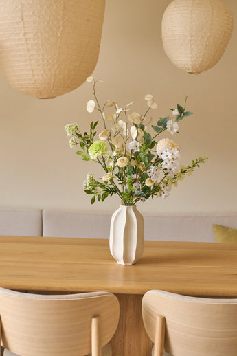 Op een houten tafel staat een witte vaas met een elegant boeket kunstbloemen van Zijden, genaamd Claire de Lune. Er staan twee beige stoelen en boven de tafel hangen papieren lantaarns. De achtergrond bestaat uit een zacht crèmekleurige muur met een bank met kussens, wat een minimalistische en gezellige sfeer creëert die doet denken aan de serene sfeer van Claire de Lune.