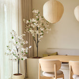 Een minimalistische eethoek met stoelen van licht hout, een ronde tafel, twee grote papieren lantaarns, doorschijnende gordijnen en kunstplanten van Kunst Kersenbloesem van 180 cm met witte kunstmatige kersenbloesembomen, die een rustige en luchtige sfeer creëren.