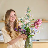 Een vrouw met lang, golvend haar schikt een boeket ‘Lavender Dreams’ van Zijden kunstbloemen in een groene vaas in haar moderne keuken, terwijl ze glimlachend aan het werk is. Het levendige boeket van zijden bloemen brengt tijdloze schoonheid in haar huis.