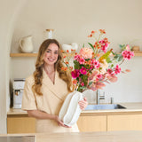 Een vrouw in een beige outfit staat in een moderne keuken. Ze glimlacht en houdt het boeket ‘Wild Peach’ van Zijden kunstbloemen vast, dat in een strakke witte vaas is geschikt en vol kleurrijke bloemen staat. Op de achtergrond zijn planken te zien met keukengerei en een koffiezetapparaat.