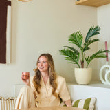Een glimlachende vrouw met lang blond haar zit in een moderne, lichte kamer met een roze kopje in haar hand. Om haar heen staan groene planten, waaronder de weelderige Kunstplant Phoenix van 85 cm, evenals houten planken en decoratieve voorwerpen.