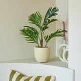 Een Kunstplant Phoenix van 85 cm in de vorm van een potpalm staat op een witte plank naast een roze kaars en een wit decoratief voorwerp. Daaronder ligt een groen-wit gestreept kussen op een bankje, terwijl op de voorgrond een deel van een houten tafel te zien is.