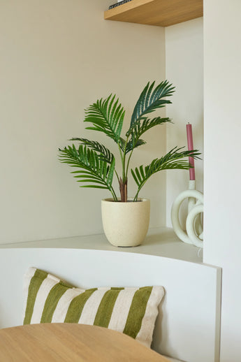 Een Kunstplant Phoenix van 85 cm in de vorm van een potpalm staat op een witte plank naast een roze kaars en een wit decoratief voorwerp. Daaronder ligt een groen-wit gestreept kussen op een bankje, terwijl op de voorgrond een deel van een houten tafel te zien is.