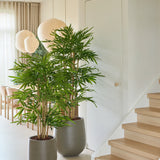 Twee Kunstplant Bamboe-potplanten van 120 cm staan naast een lichte houten trap in een licht, modern interieur met grote ramen, lichte gordijnen, ronde papieren lantaarns en een neutrale inrichting.