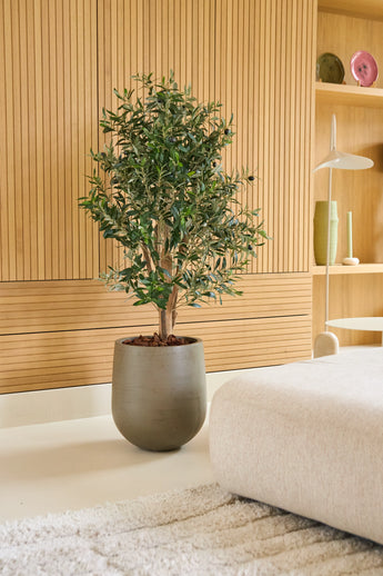 Een Kunst Olijfplant 120 cm UV staat op een lichte vloer naast een beige bank in een moderne kamer met houten lambrisering en minimalistische planken waarop vazen en servies staan uitgestald. Dit is een onderhoudsvrije olijfboom die perfect past bij een stijlvolle, onderhoudsvriendelijke inrichting.