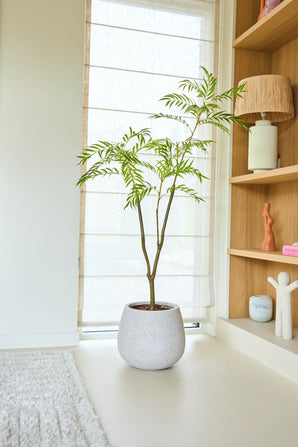 Een Kunstplant Mahonia van 150 cm met slanke groene bladeren staat op een lichte vloer bij een raam met transparante jaloezieën, naast een houten plank met decoratieve spullen en een zacht vloerkleed.