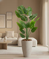 De Kunstplant Strelitzia 240 cm (incl. pot), met grote groene bladeren, staat in een moderne woonkamer met neutrale meubels, abstracte muurkunst en zachte natuurlijke verlichting.