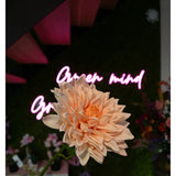 Een Kunst Georgine Real Touch lichtroze 70cm dahlia in focus, met een wazig neon "Open mind" bord en kleurrijke bloemen op de achtergrond.