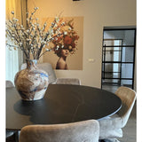 Een moderne eethoek met een ronde zwarte tafel, beige stoelen en een grote keramieken vaas met Kunst Japanse Kersenbloesem 94 cm wit. Een muurschildering van een vrouw met bloemen in haar haar siert de achtergrond.