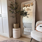 Een gezellige, moderne kinderkamer met neutrale tinten is voorzien van een golvende vloerspiegel, de Kunst Olijfboom 120 cm voor een mediterrane touch, een pluche stoel met een kussen met een berengezicht en een wieg die weerspiegeld wordt in de spiegel - de perfecte inrichting voor elke ruimte.