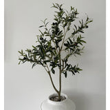 De Kunst Olijfboom 120 cm heeft groene bladeren en zwarte olijven in een witte pot met structuur en kiezels, waardoor het een prachtig decoratief element is tegen een effen lichtgrijze muur.