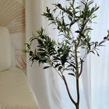 Een Kunst Olijfboom 120 cm met groene bladeren staat op een tegelvloer naast een crèmekleurige bank en geeft de kamer een Mediterrane sfeer, voor witte gordijnen en een muur met subtiel patroon.