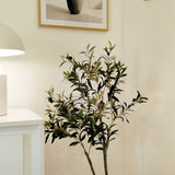 Een Kunst Olijfboom 120 cm met groene bladeren staat in de hoek van een lichte kamer en brengt een mediterraan tintje. Erboven hangen twee ingelijste kunstprints van vazen met takken, terwijl een witte kast en lamp gedeeltelijk zichtbaar zijn aan de linkerkant.