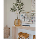Een moderne werkplek met een houten bureau, twee krukjes, een grote gouden plantenbak met de Kunst Olijfboom 120 cm voor een mediterrane flair, een wasbord met kantoorbenodigdheden en een rieten mand op de vloer als decoratief detail.