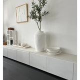 Een minimalistische kamer met een wit dressoir, de Kunst Olijfboom 120 cm als decoratief element, decoratieve witte schalen, een kaars en abstracte wandkunst. Donkerhouten vloeren en moderne accenten creëren een serene Mediterrane sfeer.