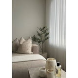 Een minimalistische woonkamer heeft een beige bank, structuurkussens, een Kunst Olijfboom 120 cm als decor, transparante witte gordijnen en een marmeren tafel met drie crèmekleurige vazen en parfum. Natuurlijk licht verlicht de rustige ruimte.