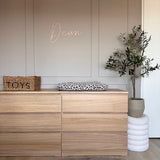 Een minimalistische kinderkamer bevat een houten commode met een aankleedkussen met luipaardprint, een geweven mand met het label "Toys" als decoratie, de Kunst Olijfboom 120 cm voor groen en "Dean" in cursief op de beige muur erboven.