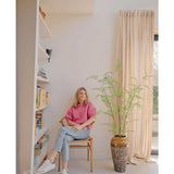 Een vrouw in een roze trui en spijkerbroek zit op een houten stoel naast de Kunst varen plant 140 cm, in de buurt van een boekenplank en een groot raam met beige gordijnen in een lichte, moderne woonruimte.