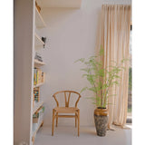 Een minimalistische kamer heeft een houten stoel naast een Kunst varen plant van 140 cm, transparante gordijnen voor het raam en boekenplanken met boeken en decor tegen een witte muur.