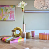 Een moderne houten tafel toont kleurrijke objecten, waaronder een roze vaas met daarin de Kunstbloem Amaryllis Real Touch wit 66 cm, een gele ringsculptuur, gestreepte boeken en een roze beker. Abstracte kunst accentueert de achtergrondmuur.