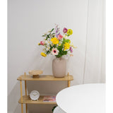Een lichthouten plank toont een beige vaas met Kunstranonkel Real Touch Geel 60cm bloemen, een witte klok en een houten dienblad. Een deel van een ronde witte tafel verschijnt op de voorgrond tegen een effen witte muur.