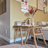 Een moderne woonkamerhoek met een lichte houten salontafel met daarop de Kunstbloem Anemoon vertakt 56 cm roze in een roze vaas, samen met een lamp en boeken. Ernaast staat een beige bank, geaccentueerd door muurkunst en een galerieachtig fotobord op de gele muur.