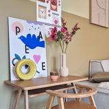 Een houten bureau met een bijpassende stoel toont een blauwe vogelafdruk, Kunstbloem Anemoon vertakt 56 cm roze, een rond geel ornament en een klein kopje. Boven hangt een prikbord met foto's en notities naast een beige bank.