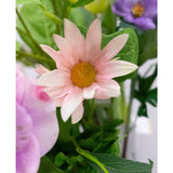 Een close-up van de Kunstbloem Aster 60 cm roze bloemblaadjes met een geel hart, omringd door groene bladeren en andere bloemen, waaronder een paarse, op de achtergrond.
