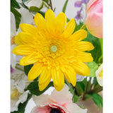 De Kunstbloem Gerbera 53 cm geel, een levendige gele kunstbloem met gelaagde bloemblaadjes, staat tussen andere kleurrijke kunstbloemen en groene bladeren, met witte en roze bloemen op de achtergrond.
