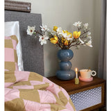 Een nachtkastje met een blauwe vaas met daarin de Kunstbloem Magnolia 50 cm geel/oranje, een roze theekopje op een margrietvormig schoteltje en een brandende kaars staat naast een bed dat is bedekt met een roze en bruine deken met geometrische patronen.