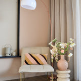 Een moderne, gezellige hoek met een crèmekleurige stoel met een gestreept kussen, een hoge boogvormige vloerlamp, beige gordijnen, een houten vloer en een ronde vaas met Kunstbloem Fritillary 94 cm wit bloemen op een gebeeldhouwde bijzettafel.