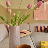 Een Kunstbloem Franse Tulp van 63 cm in roze staat in een vaas op de tafel naast een oranje kopje in een gezellige woonkamer met een crèmekleurige bank, kussens met patronen, een gele krat en abstracte wandkunst op de achtergrond.