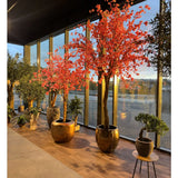Potplanten en -bomen, zoals de Kunstboom Kersenbloesem roze van 330 cm met felroze bloesems, staan opgesteld bij hoge glazen ramen die het licht op de houten vloer weerkaatsen.