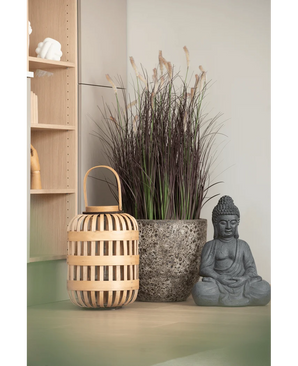 Een houten lantaarn, een hoge stenen pot met Kunstgras-uiengras-bonsai van 100 cm (brandvertragend) en een grijs Boeddhabeeld staan opgesteld op een lichtgekleurd oppervlak, vlakbij een houten plank en een bleke muur.