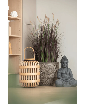 Een houten lantaarn, een hoge stenen pot met Kunstgras-uiengras-bonsai van 100 cm (brandvertragend) en een grijs Boeddhabeeld staan opgesteld op een lichtgekleurd oppervlak, vlakbij een houten plank en een bleke muur.