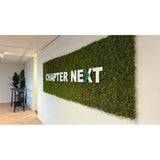 Een luxe interieuraccent: een groene wanddisplay met Kunsthaag Nordic Mos 50x50 cm UV-kaders, met "CHAPTER NEXT" in vette 3D-letters. Een tafel versierd met planten maakt het geraffineerde tafereel compleet.