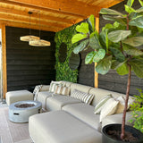 Een gezellige zithoek buiten met een beige sofa, gestreepte kussens, een ronde vuurplaats, een grote plant in potten en een houten pergola. De muur is voorzien van groen met de Kunsthaag Volta 100x100 cm UV voor extra stijl.