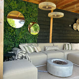 Een gezellig terras met een beige sofa, gestreepte kussens, vuurplaats en geweven vloerkleden. Aan de muur hangt UV-groen van Kunsthaag Volta 100x100 cm, ronde spiegels en rieten lampen onder een houten pergola.
