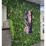 Een wand bekleed met Kunsthaag mat Savanne 100x100 cm, met UV-beschermd groen in een kunststof plantenbak Ø100, heeft een groot zwart-wit portret in het midden. Het moderne interieur heeft een betegeld plafond en een houten balie aan de linkerkant.