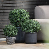 Drie ronde, groene Kunst Buxus op stam UV d30 cm struiken in geribbelde zwarte potten staan op een houten vlonder naast een lichtgrijze bank, met daarachter een donkerhouten muur.
