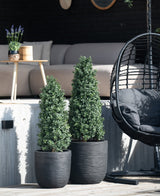 Twee Kunstplant Buxus Pyramide UV 90 cm vormbomen staan op een patio bij een buitenbank en een ronde rieten hangstoel met een zwart kussen, terwijl een klein tafeltje met een plant op de achtergrond staat.