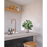 Een modern aanrecht met een messing kraan, twee zeepdispensers, de Kunstplant Tabaksplant Fiddle Leaf UV 45 cm potplant en twee houten krukjes. Een zwevende plank met potten en flessen is gemonteerd op een lichtgekleurde muur.