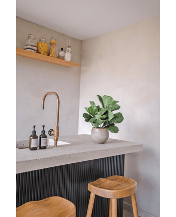 Een minimalistisch aanrecht met de Kunstplant Tabaksplant Fiddle Leaf 45 cm, twee houten krukjes, een gouden kraan, zeepdispensers en een zwevende plank met ingrediëntenpotjes tegen een lichtgekleurde muur.
