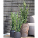 Een Kunstgras Groen UV 90cm kunstplant in een zwarte pot staat op een houten vloer naast een grijze buitenbank, met een donkere houten lattenwand op de achtergrond.