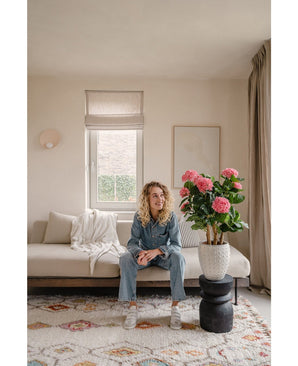 Een vrouw in een spijkerpak en witte sneakers zit op een beige bank in een lichte woonkamer. Naast haar staat de Kunstplant Hortensia 85 cm roze, die roze bloemen en groen toevoegt aan de lichte ruimte.