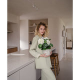 Een vrouw met krullend blond haar, gekleed in een lichtgroene outfit, staat in een moderne keuken met een Kunstplant Hortensia Wit UV 35 cm in haar handen – met crèmewitte bloemen – en glimlacht naar de camera.