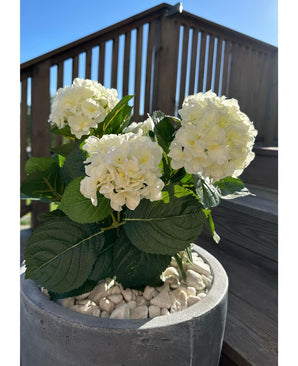 De Kunstplant Hortensia Wit UV 35 cm, met witte bloemen en groene bladeren, staat op een zonnige dag buiten op witte kiezelstenen, met op de achtergrond houten terrasleuningen en een trap.